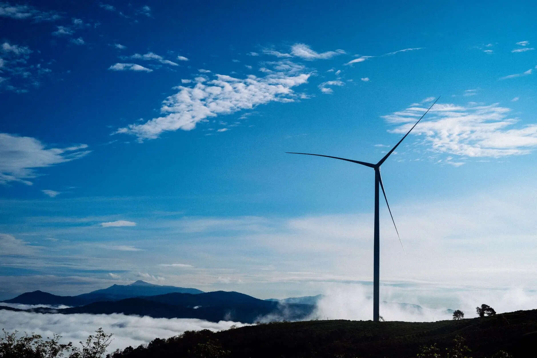 A solitary wind turbine rises against a vibrant blue sky, surrounded by clouds and distant mountains.